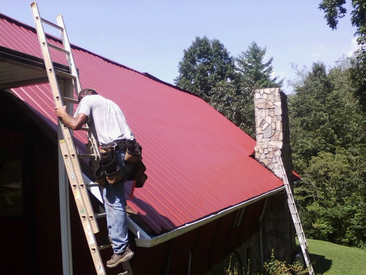 Roofer on a ladder beside a red metal roof and stone chimney — Soffit & Fascia Repair by Roofing Brothers in Frackville